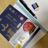 机の上にノートを広げ、その上に本とペンが置かれている写真